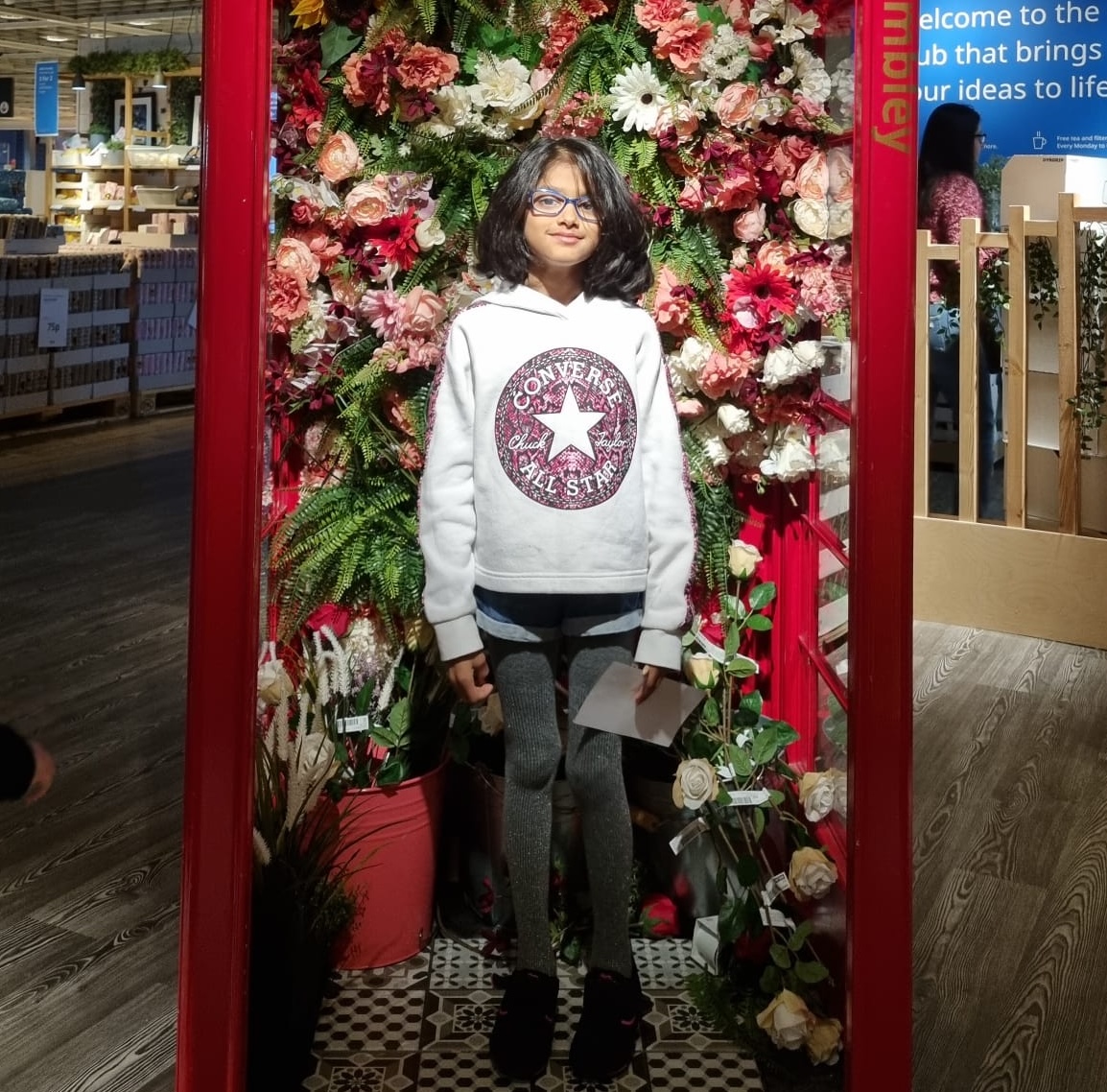 Riya is standing in a converted red phone box and surrounded by red, white and pink flowers